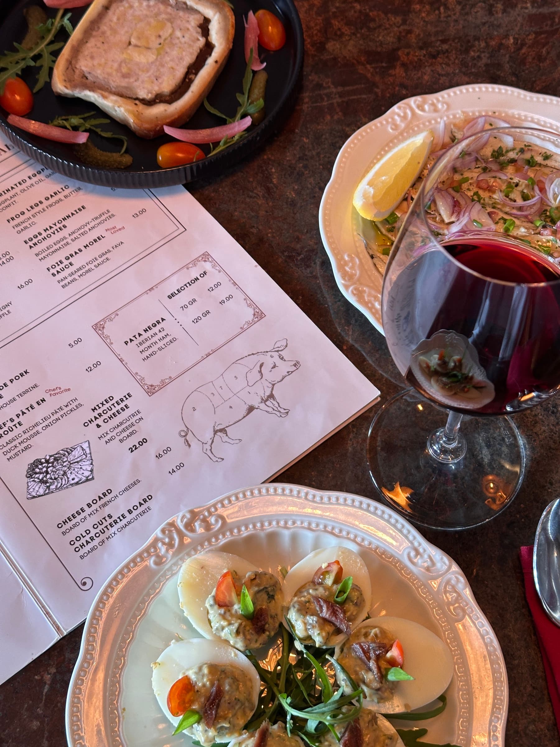 An overhead shot of Somaha's charcuterie menu with a hand-drawn pig cut diagram, a glass of red wine being poured, and deviled eggs