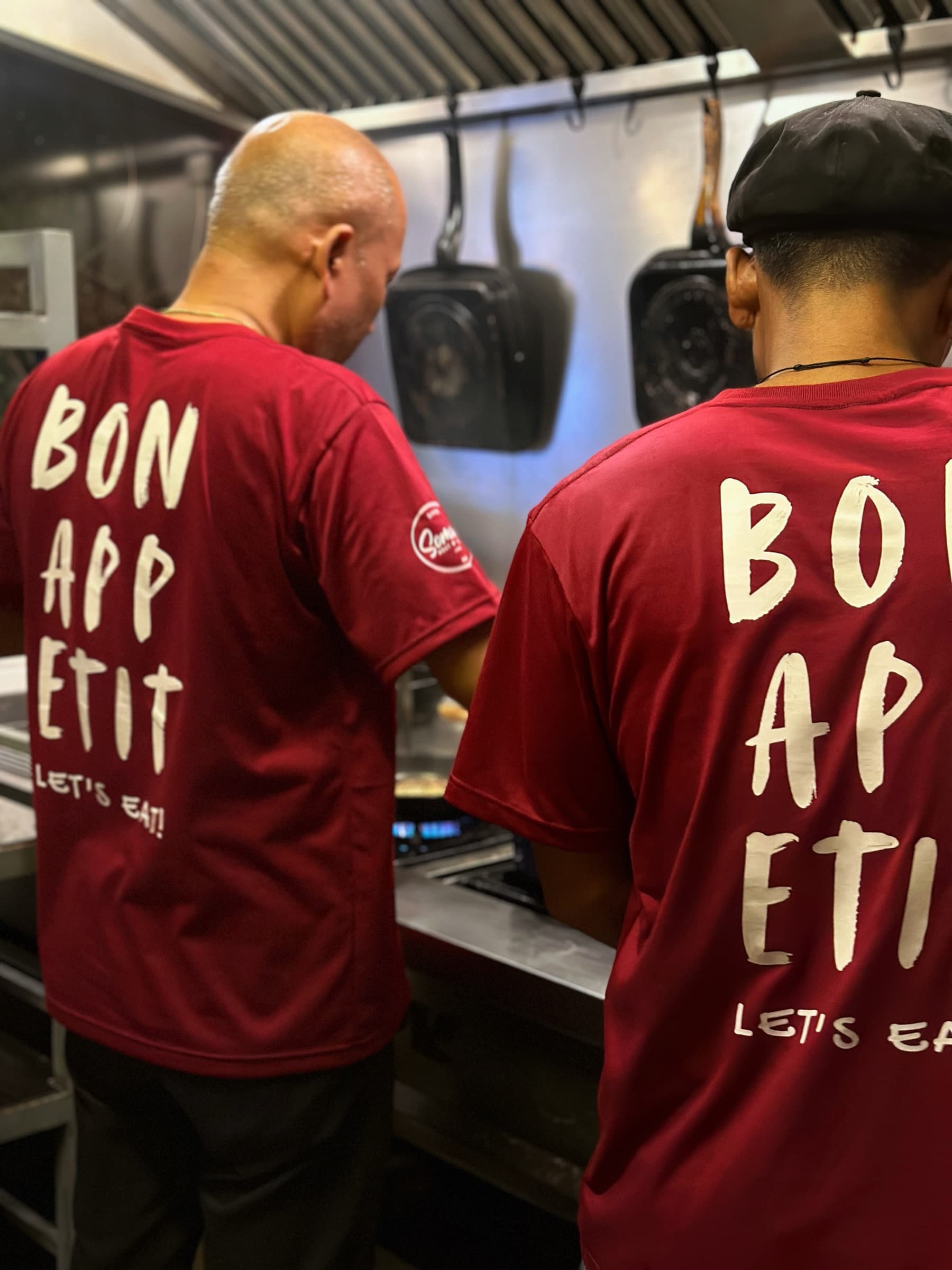 Two Somaha cooks, backs turned, in “Bon Appétit — Let's Eat!” kitchen t-shirts working the line